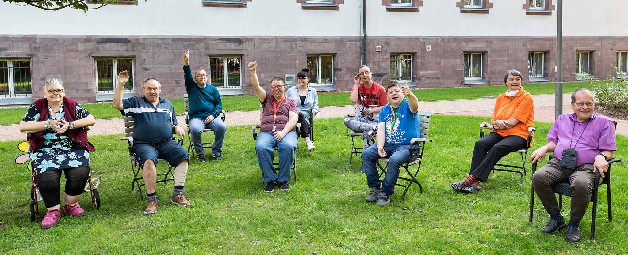 Klienten der Voithea sitzen auf Stühlen im Garten und grüßen.